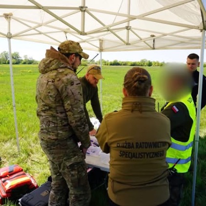 Grupa osób pod białym namiotem ekspozycyjnym na otwartym polu. Wśród nich są osoby w mundurach kamuflażowych oraz w bluzach z napisem „SŁUŻBA RATOWNICTWA SPECJALISTYCZNEGO”. Analizują wspólnie dokumenty rozłożone na stole.