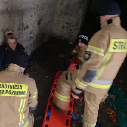Akcja ratunkowa w ciemnym, betonowym pomieszczeniu. Strażacy w piaskowych mundurach z napisem "STRAŻ" przygotowują pomarańczowe noszy typu deska, aby pomóc osobie siedzącej pod ścianą.