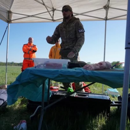 Mężczyzna w mundurze kamuflażowym prowadzi szkolenie pod namiotem na świeżym powietrzu. Na stole przed nim leżą materiały medyczne oraz trenażer medyczny imitujący ranę na kończynie. W tle widać inne osoby w jaskrawych strojach ratowniczych.