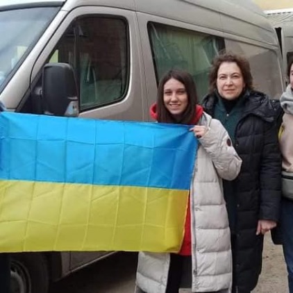 Dwie kobiety stoją przed srebrnym samochodem dostawczym i trzymają przed sobą rozłożoną flagę Ukrainy (niebiesko-żółtą). Kobiety są ubrane w zimowe kurtki i uśmiechają się do aparatu.